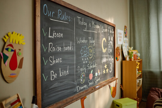 Kindergarten classroom chalkboard displaying daily schedule, weather chart, and classroom rules with colorful drawings, educational content, and creative decorations visible on wall