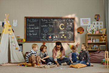 Multiethnic group of children sitting on floor with young adult Black man teacher exploring flashcards with alphabet letters in kindergarten classroom, engaging in educational activity near chalkboard © Seventyfour