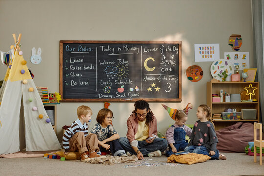 Multiethnic group of children sitting on floor with young adult Black man teacher exploring flashcards with alphabet letters in kindergarten classroom, engaging in educational activity near chalkboard