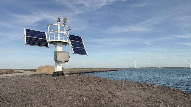 Complete weather station with solar panels and anemometer in coastal landscape setting, blue sky with clouds, multiple wind turbines visible across water, Dutch environmental monitoring.