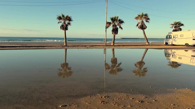 Stunning scenic view of calm ocean waves breaking on sandy beach with palm trees and a camper van, reflected in a large puddle under clear blue sky