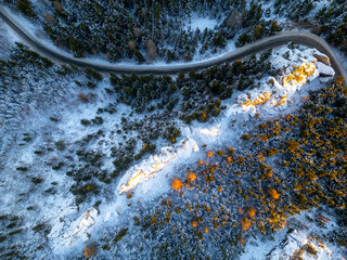 Fototapeta premium A view from above shows a rock formation covered in snow with a winding road nearby. Trees surround the area, showing hints of winter colors in the landscape.