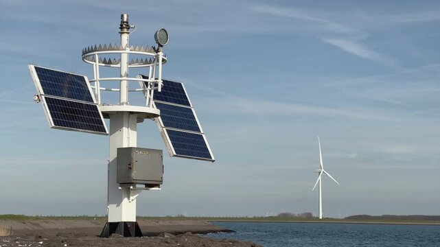 Detailed close-up view of coastal weather station featuring solar panels, anemometer and monitoring equipment, single wind turbine visible in background across water.