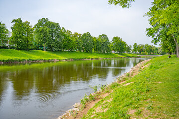 Fototapeta premium Jirsakovy sady in Hradec Kralova shows green trees along the riverside. People are walking and sitting on the grass. The Elbe and Orlice rivers meet in this area.