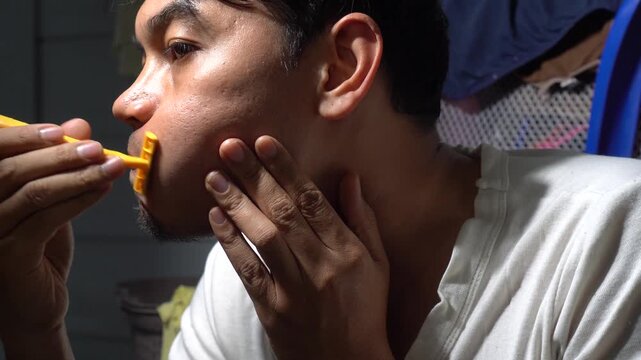 Close up of a middle-aged Southeast Asian man shaving his facial hair using a yellow disposable razor. Authentic grooming routine, male skincare, and morning hygiene concept in a domestic setting with
