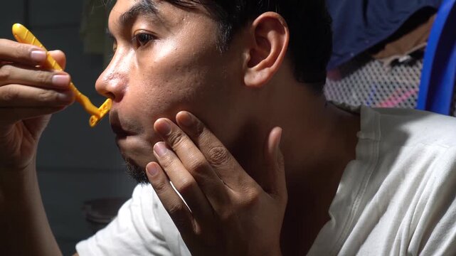 Close up of an Asian man shaving his mustache and facial hair with a manual yellow razor at home. Focused expression during morning grooming routine and personal hygiene. Real skin texture with lighti