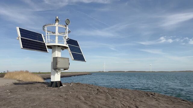 Weather station with solar panels and anemometer against dramatic sky with clouds, multiple wind turbines visible across water in background.
