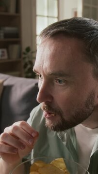 Vertical close-up of face of 30-year-old Caucasian man attentively watching horror movie or slasher flick on TV, eating chips, displaying vivid emotions of fear, dread, shock at violent content