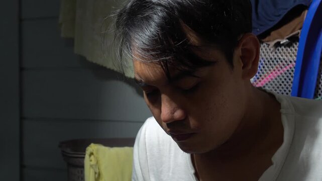 An Asian man in a white shirt trimming his beard and neck hair using a cordless electric clipper. Natural indoor lighting focusing on self-grooming at home. Concept of DIY haircut and maintaining a cl