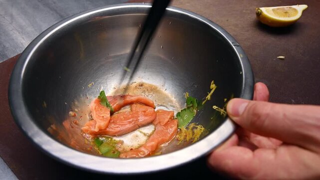 Hand using chopsticks to mix salmon slices with herbs in metal bowl