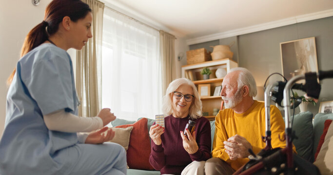 A home health nurse sits with an elderly couple in their living room, discussing their medication. The couple is engaged and attentive as they hold medicine bottles.
