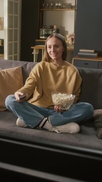 Full vertical shot of happy, relaxed Caucasian woman sitting comfortably on sofa with crossed legs with bowl of popcorn, switching TV channels, searching for fun movie or sitcom on streaming service