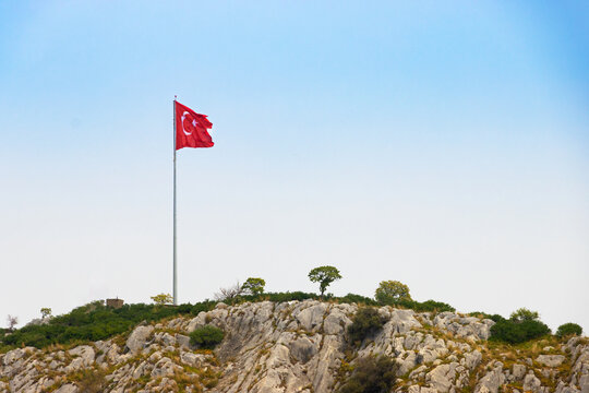 Waving Turkish national flag on a high flagpole at the top of a steep rocky hill against a clear blue sky, panoramic view of a mountain summit for travel and national concepts.