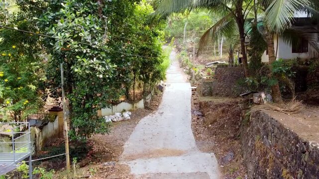 A steady, high-angle shot of a narrow concrete path winding through a lush, tropical hillside village, featuring vibrant greenery, palm trees, and traditional stone walls.