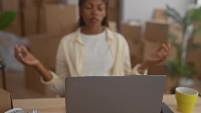 Woman meditating with hands in mudra at laptop amid moving boxes in a building; mindfulness calm transition.