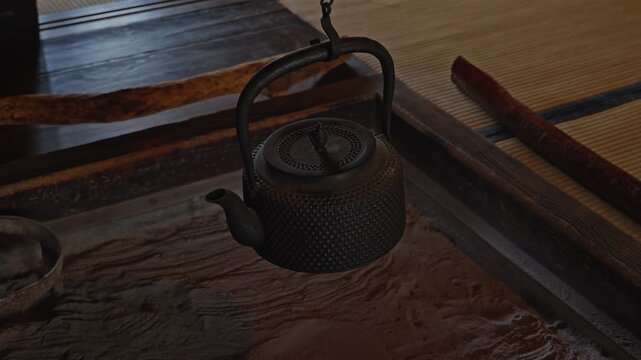 A view of a traditional Japanese cast iron kettle (tetsubin) suspended by a chain over a charcoal hearth (irori) inside a classic minka home with tatami matting.
