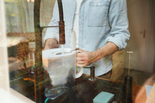 Man filling water filter jug with tap water in kitchen at home