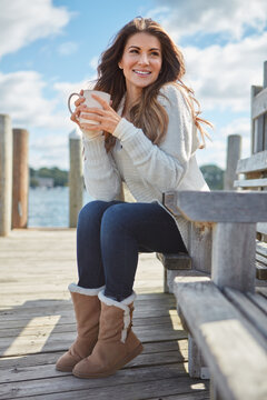 Woman, thinking and coffee on pier with lake, relax and contemplation for peace on winter vacation. Person, smile outdoor and cold weather on bench with river, warm beverage and travel reflection.