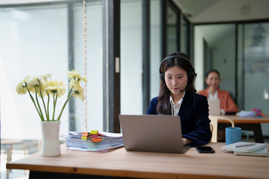 Young Asian woman wearing a headset and working intensely on a laptop, actively engaging with clients and managing tasks while providing remote support in a shared office environment