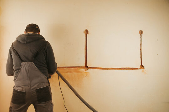 A worker using a wall chaser machine to cut grooves in a wall for electrical installations. High-quality construction photography showing professional power tools and renovation process indoors.