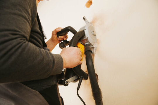 A worker using a wall chaser machine to cut grooves in a wall for electrical installations. High-quality construction photography showing professional power tools and renovation process indoors.