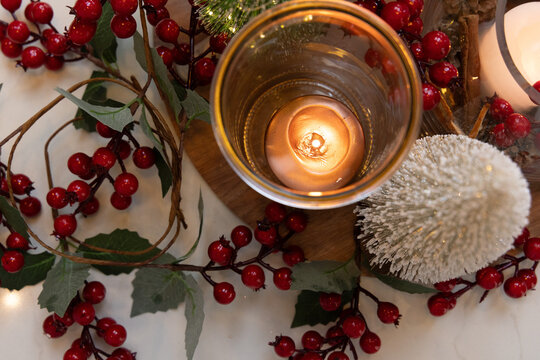 Glowing cylindrical glass votive on wooden board on marble table, holly berries and glittered tree