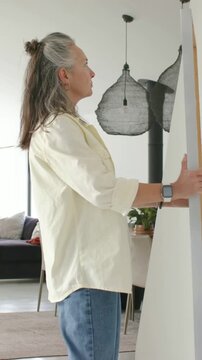 Vertical video: Carrying senior entering door, peeking then hanging canvas on white wall in yellow
