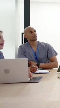 Vertical video: Leaning two staff in scrubs at table, gesturing, glancing at screen, writing notes