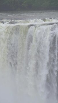 slow motion video of water fall crashes over a steep cliff. Rushing torrents form a frothy, white curtain. Green trees frame the distant, misty horizon. Powerful nature dominates the scene with raw