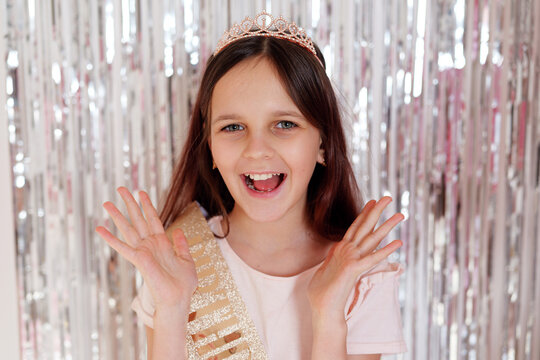 A girl with a tiara and sash smiles widely while standing in front of shiny curtains. She raises her hands in excitement during a fun celebration