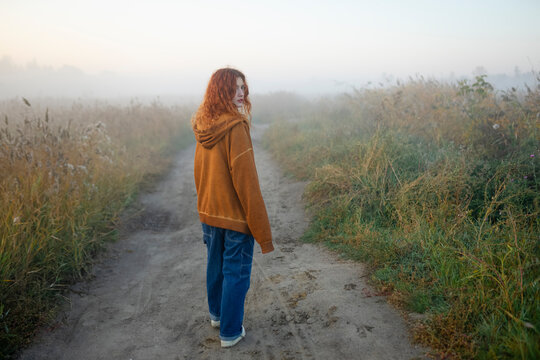 Woman with red hair in orange hoodie walking on foggy park path