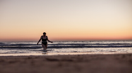 Obraz premium Body positivity: A girl emerges from the sea after swimming and enjoys a beautiful sunset on the beach. Summer mood.