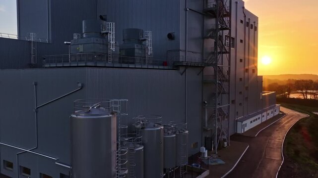 Aerial drone rising shot above large modern industrial facility in Latvia at sunset with sun low on horizon illuminating gray buildings cooling towers silos ladders piping and rural landscape