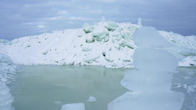 Pale meltwater pool beside a jagged sea-ice ridge under a cloudy sky, with translucent ice blocks in the foreground. Cold winter seascape showing fragile thaw conditions.