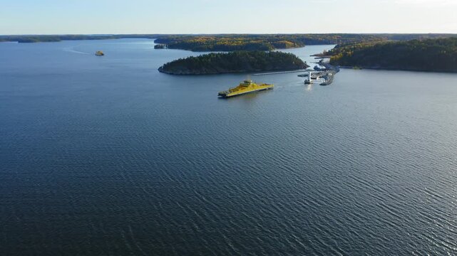 Yellow open deck hybrid island car ferry making way ahead in Finnish archipelago. High altitude aerial view. Vessel shortly departed from the loading ramp area.