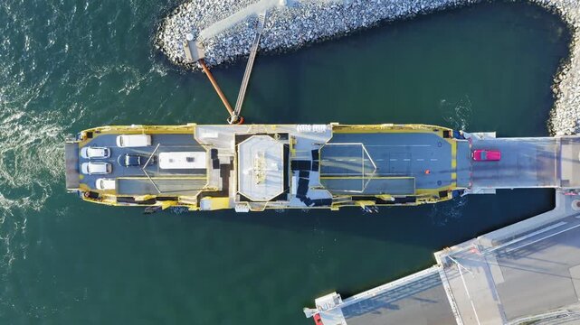 Yellow open deck island car ferry loading vehicles from an end of road loading ramp in Finnish archipelago. Aerial top down view. Vessel using propulsion to stay in position for loading.
