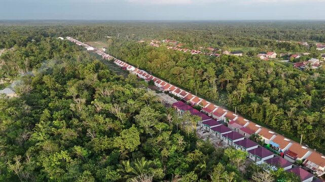 A drone shot moves forward, sweeping past a long row of new houses. Colorful rooftops stretch out amid a lush green forest under the soft sunlight. 