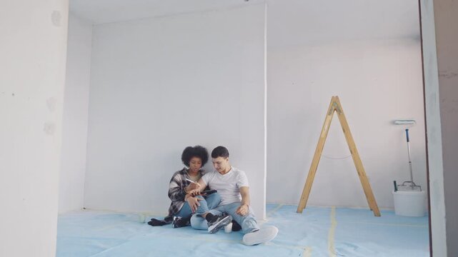 A man and a woman sit on a protective blue cover in a room under renovation, sharing a light moment. Unfinished walls and a wooden ladder suggest ongoing home improvement, with paint trays nearby.