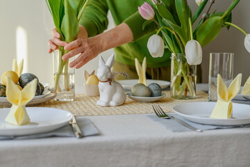 Woman decorating table for Easter holiday dinner. Person hands put flowers in vase on table with aesthetic spring table setting. Table with linen cloth, plate with dyed eggs, rabbit and flowers