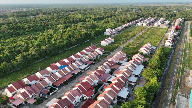 Aerial cinematic drone moves forward over rows of red-roof houses, a neat residential area stretching through lush green forest under soft daylight.