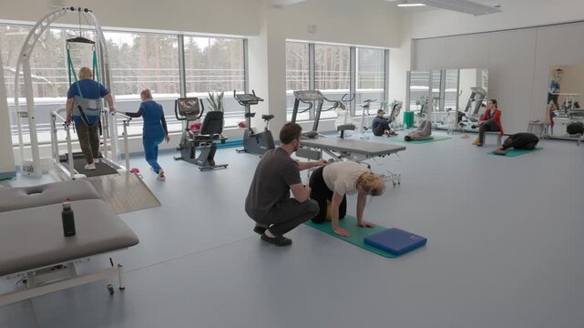 Patients performing guided rehabilitation exercises with therapists using gym and physiotherapy equipment in a bright clinical setting with large windows