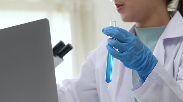Young Asian women scientist working with test tube in laboratory. Scientist in a lab coat and safety glasses handles a test tube at a desk in a laboratory, make note on laptop, experimentation 