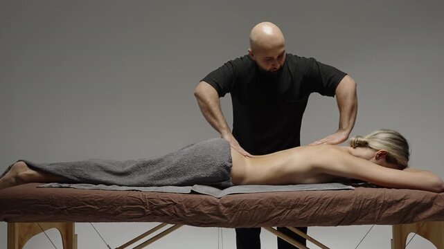Professional massage therapist performing back treatment on a client lying prone on a spa table, transitioning from hand warming to deep pressure techniques in a studio setting