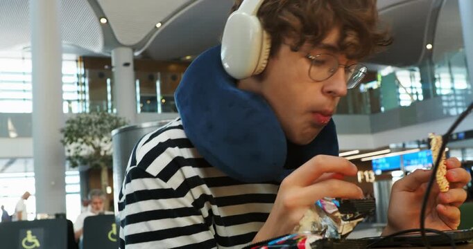 Young man with headphones and neck pillow eats a snack while waiting for his flight at an airport terminal. A candid close-up of a teen traveler relaxing and snacking in a modern departure lounge