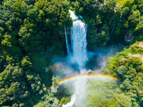 Aerial view of the cascading Marmore Falls plunges amidst verdant foliage, a vibrant rainbow arches across the misty spray, Terni, Umbria, Italy.