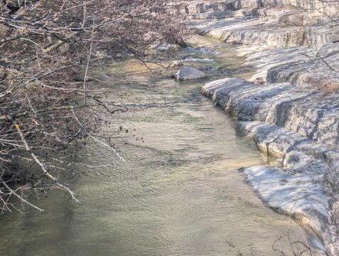 Ruscello poco profondo che scorre tra rocce stratificate, con rami spogli sul bordo e acqua limpida in movimento