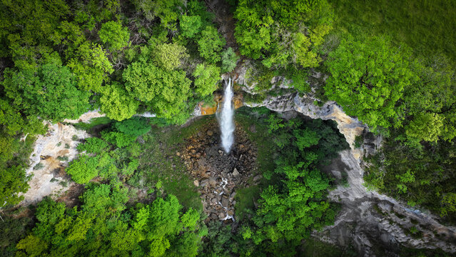 Aerial view of a waterfall cascading down a rocky cliff face, framed by lush green trees, a serene spectacle from above, Saint-Claude, Bourgogne-Franche-Comte, France.