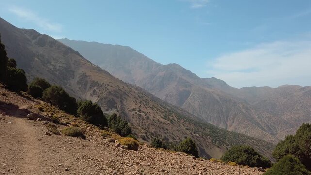 Panoramic view of rugged mountain slopes with scattered juniper trees in the High Atlas Mountains near Imlil, Morocco, beneath a clear blue sky in the scenic Mount Toubkal region.