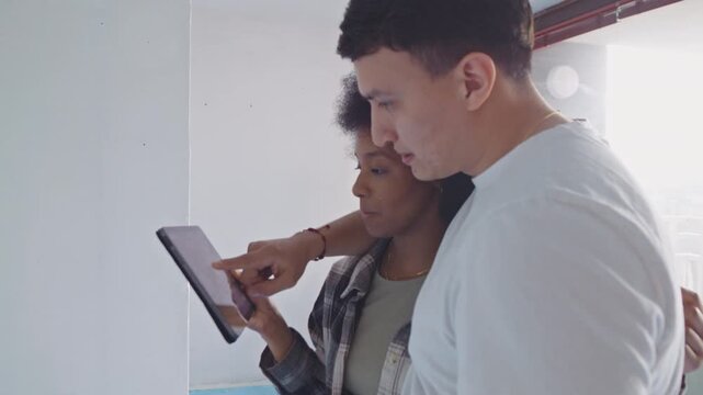 A man and woman examine a tablet displaying a room layout in an unfinished apartment, discussing renovation plans. Their focused expressions suggest detailed planning for home improvement.