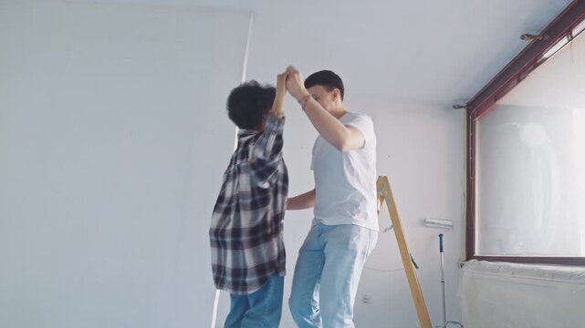 In a brightly lit room under renovation, two young men engage in playful interaction, one steadying the other amid construction materials like a step ladder and drop cloth.